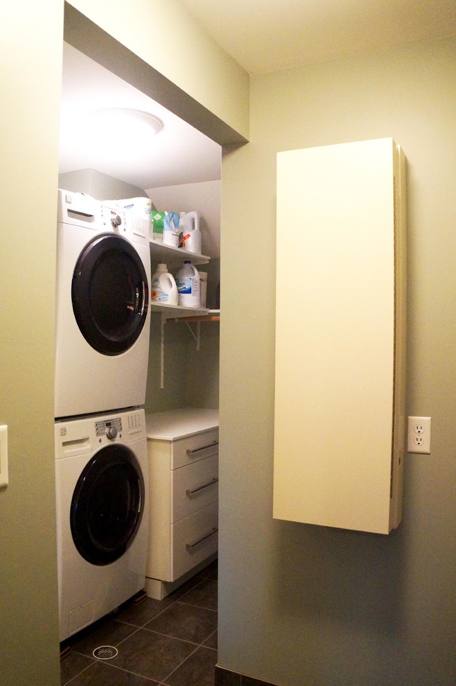 Inspiration for a mid-sized traditional galley dedicated laundry room in Detroit with flat-panel cabinets, white cabinets, solid surface benchtops, green walls, porcelain floors, a stacked washer and dryer, brown floor and white benchtop.