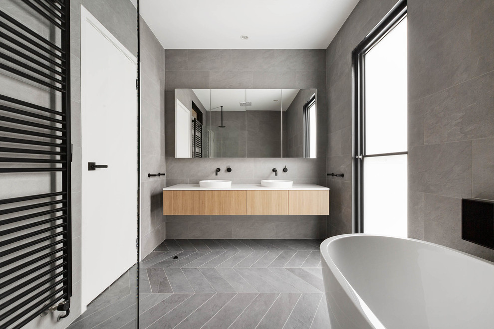 Inspiration for a contemporary bathroom in Melbourne with flat-panel cabinets, light wood cabinets, a freestanding tub, gray tile, grey walls, a vessel sink, grey floor and white benchtops.