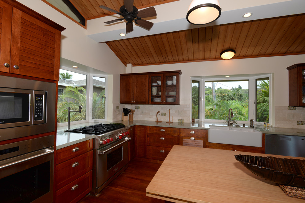 Design ideas for a large tropical u-shaped kitchen in San Diego with a farmhouse sink, beige splashback, stone tile splashback, stainless steel appliances, dark hardwood floors, brown floor, shaker cabinets, medium wood cabinets, solid surface benchtops and with island.