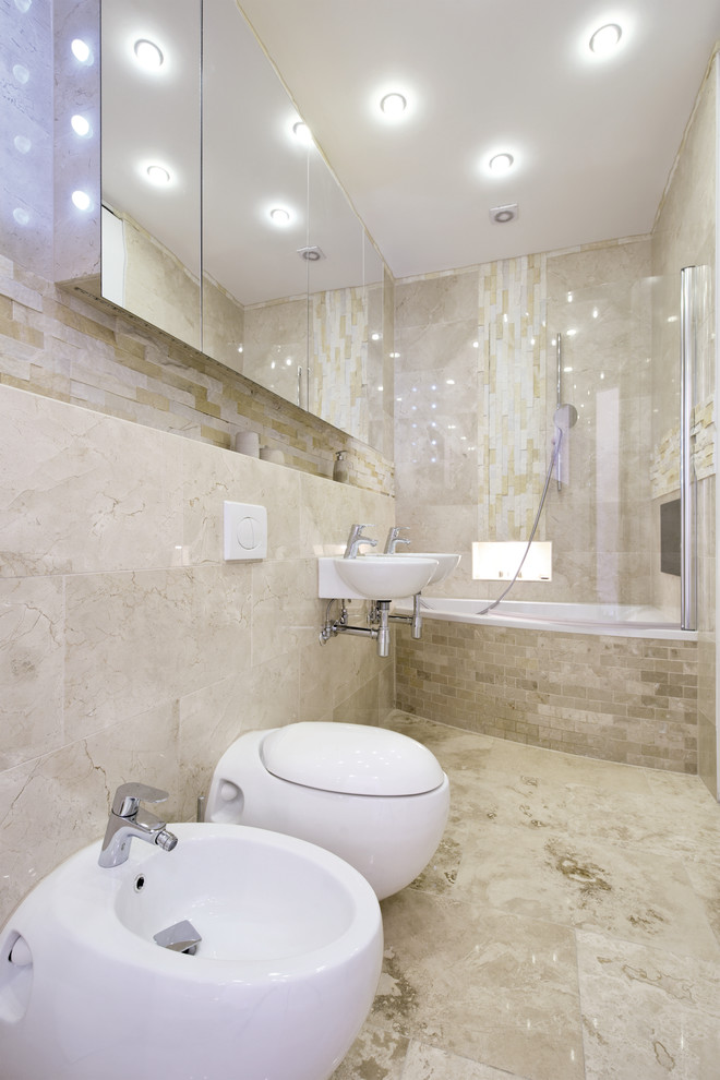 Inspiration for a mid-sized contemporary kids bathroom in London with a shower/bathtub combo, beige walls, beige floor, a hinged shower door, glass-front cabinets, a drop-in tub, a wall-mount toilet, beige tile, limestone, limestone floors and a wall-mount sink.