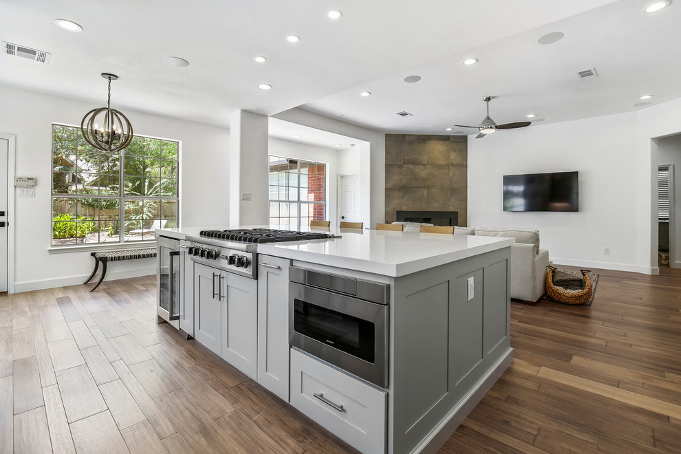 This is an example of a mid-sized transitional l-shaped eat-in kitchen in Houston with a farmhouse sink, shaker cabinets, grey cabinets, quartzite benchtops, blue splashback, porcelain splashback, stainless steel appliances, light hardwood floors, with island, brown floor and white benchtop.