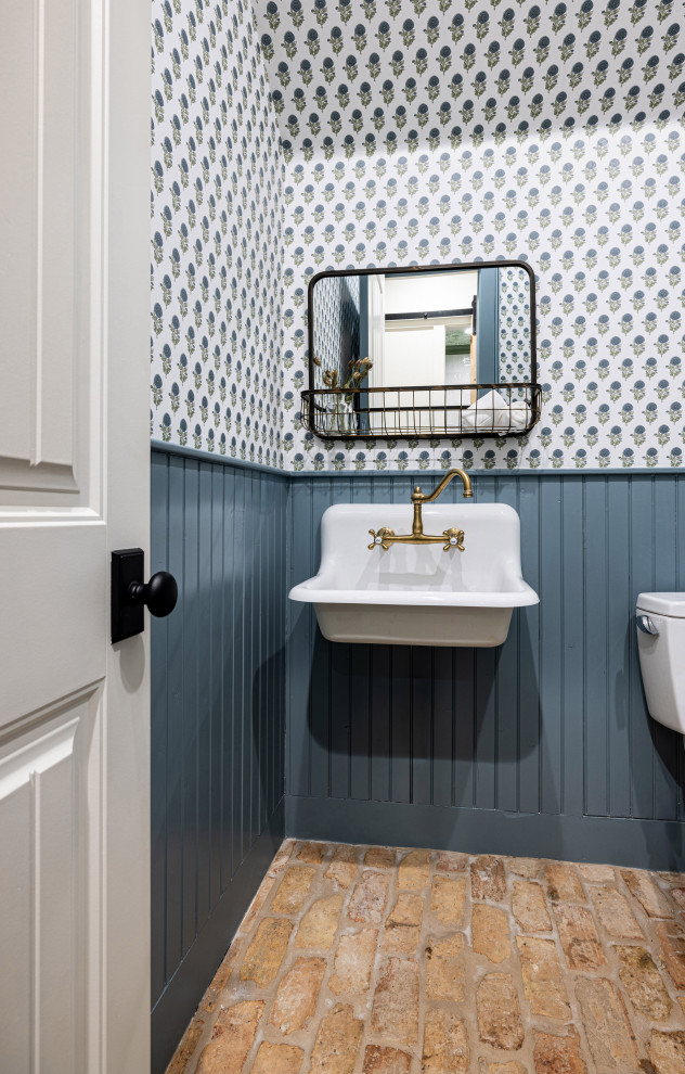 Design ideas for a small farmhouse cloakroom in Dallas with a one-piece toilet, blue walls, brick flooring, a wall-mounted sink, brown floors, a wallpapered ceiling and wainscoting.