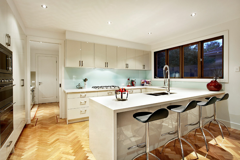 Photo of a large contemporary u-shaped open plan kitchen in Melbourne with a drop-in sink, white cabinets, quartz benchtops, blue splashback, glass sheet splashback, stainless steel appliances, medium hardwood floors and no island.