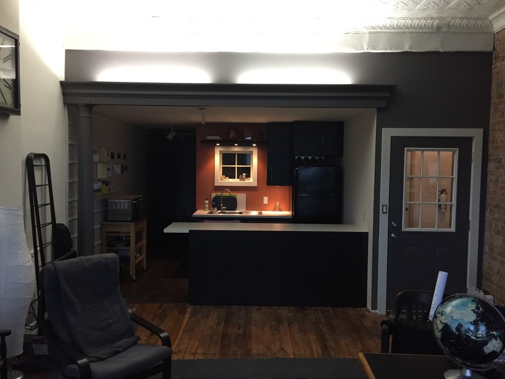 This is an example of a small industrial galley eat-in kitchen in New York with a double-bowl sink, flat-panel cabinets, black cabinets, laminate benchtops, orange splashback, black appliances, vinyl floors, a peninsula and brown floor.