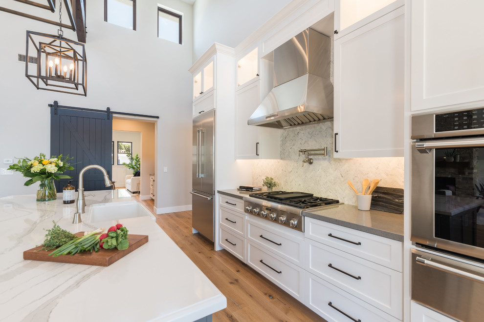 Design ideas for a mid-sized country galley eat-in kitchen in San Luis Obispo with a farmhouse sink, recessed-panel cabinets, white cabinets, marble benchtops, white splashback, stone tile splashback, stainless steel appliances, with island, white benchtop, light hardwood floors and beige floor.