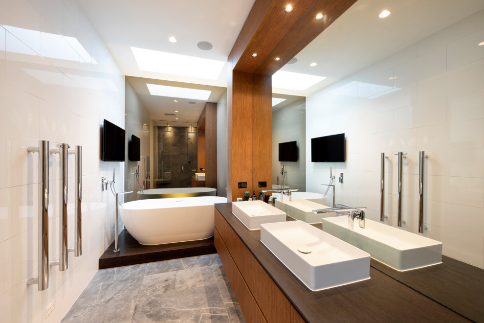 This is an example of a large contemporary master bathroom in Melbourne with flat-panel cabinets, medium wood cabinets, a freestanding tub, a double shower, white tile, porcelain tile, limestone floors, a trough sink, a hinged shower door, a double vanity and a floating vanity.