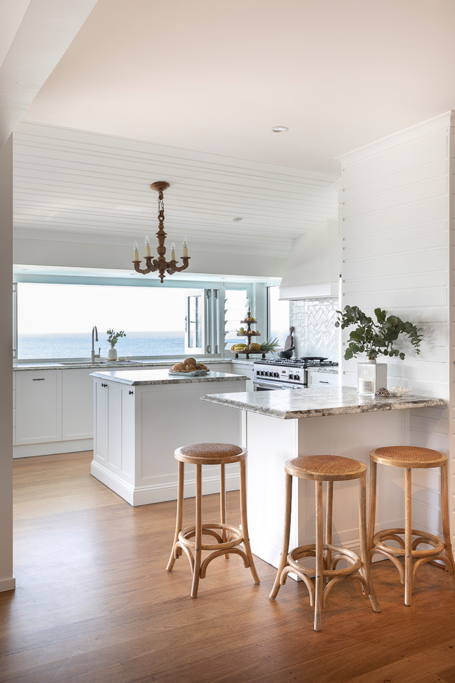 Inspiration for a mid-sized tropical u-shaped kitchen in Sunshine Coast with an undermount sink, shaker cabinets, white cabinets, granite benchtops, white splashback, ceramic splashback, stainless steel appliances, medium hardwood floors, with island, multi-coloured benchtop and brown floor.