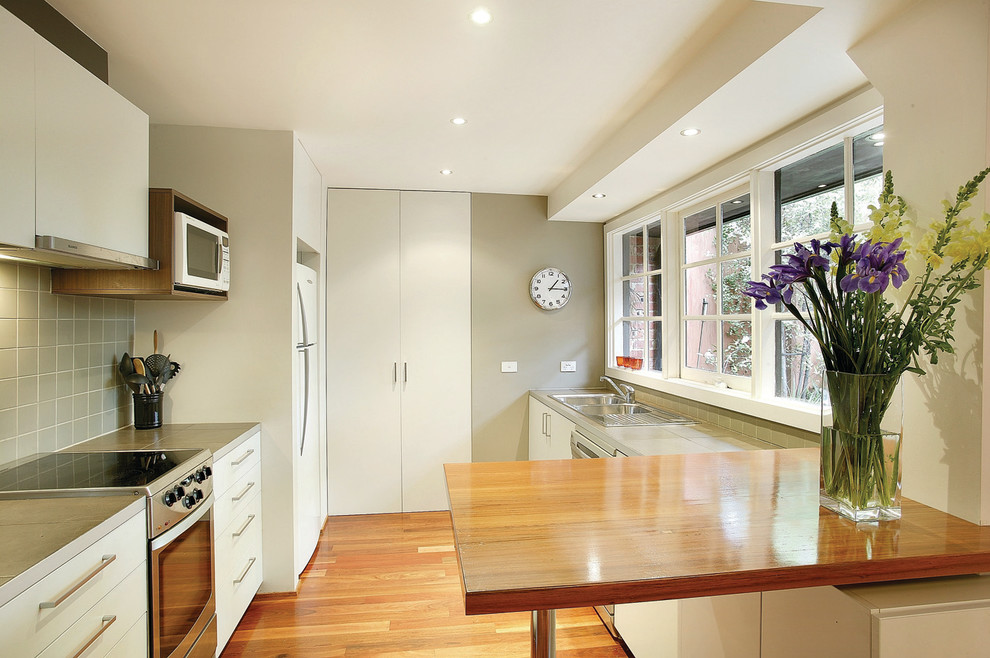 This is an example of a transitional galley separate kitchen in Melbourne with flat-panel cabinets, white appliances, wood benchtops, a drop-in sink, white cabinets and grey splashback.
