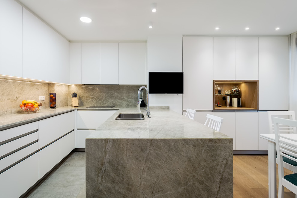 Design ideas for a mid-sized contemporary l-shaped open plan kitchen in Bilbao with a double-bowl sink, flat-panel cabinets, white cabinets, limestone benchtops, beige splashback, limestone splashback, white appliances, light hardwood floors, a peninsula and beige benchtop.