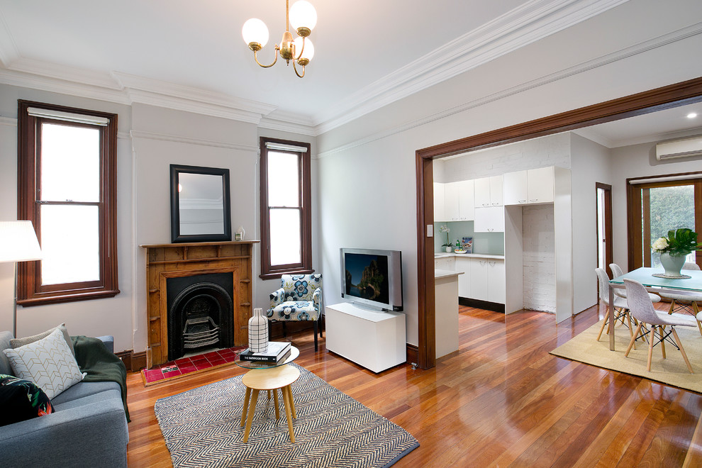 This is an example of a traditional open concept living room in Sydney with white walls, medium hardwood floors, a standard fireplace, a wood fireplace surround, a freestanding tv and brown floor.