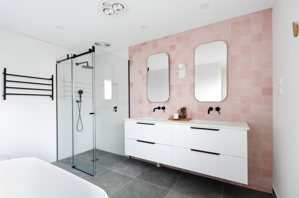 На фото: большая главная ванная комната в современном стиле с фасадами с декоративным кантом, белыми фасадами, отдельно стоящей ванной, угловым душем, унитазом-моноблоком, розовой плиткой, керамогранитной плиткой, розовыми стенами, полом из керамогранита, врезной раковиной, столешницей из искусственного кварца, серым полом, душем с раздвижными дверями, белой столешницей, фартуком, тумбой под две раковины и подвесной тумбой с