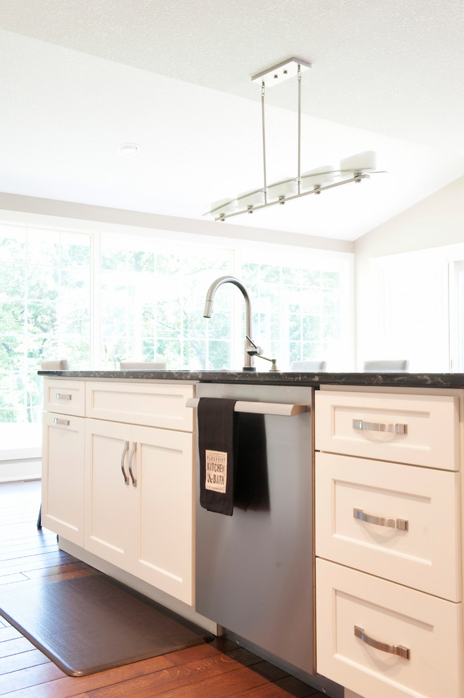 This is an example of a large transitional l-shaped eat-in kitchen in Other with an undermount sink, recessed-panel cabinets, white cabinets, quartz benchtops, beige splashback, limestone splashback, stainless steel appliances, medium hardwood floors, with island, brown floor and black benchtop.