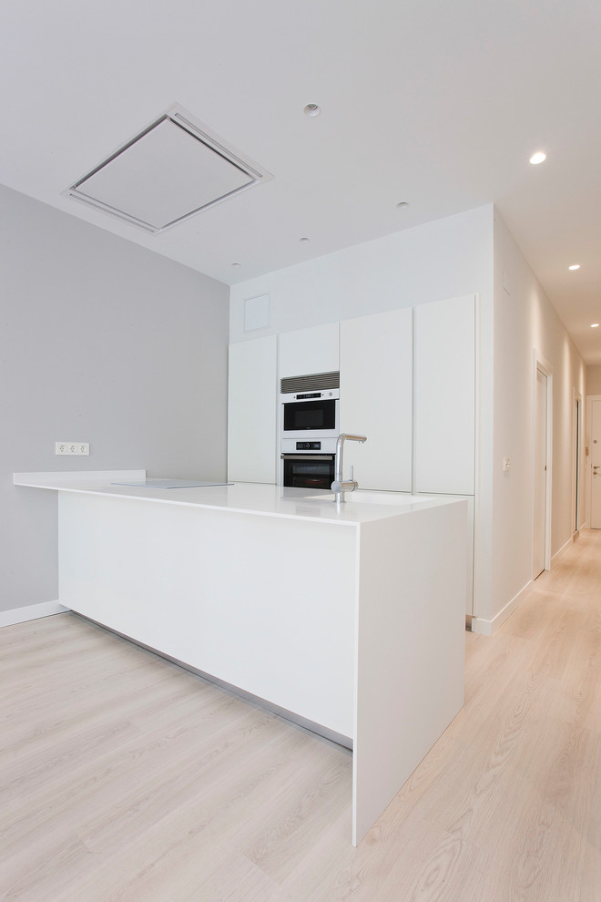 Photo of a mid-sized contemporary single-wall open plan kitchen in Other with an integrated sink, flat-panel cabinets, white cabinets, white appliances, light hardwood floors, a peninsula and beige floor.