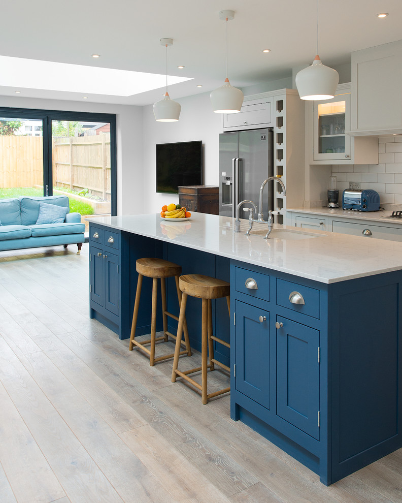 Design ideas for a mid-sized transitional eat-in kitchen in London with an undermount sink, shaker cabinets, blue cabinets, white splashback, subway tile splashback, stainless steel appliances, light hardwood floors and with island.