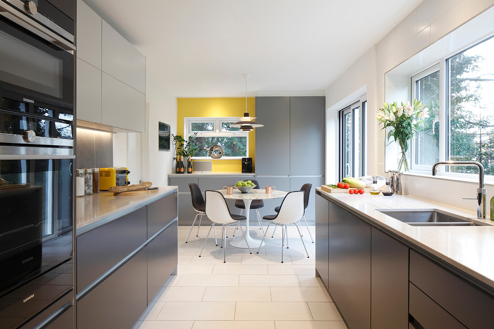 Contemporary galley eat-in kitchen in London with flat-panel cabinets, grey cabinets, solid surface benchtops, grey splashback, black appliances, ceramic floors and a double-bowl sink.