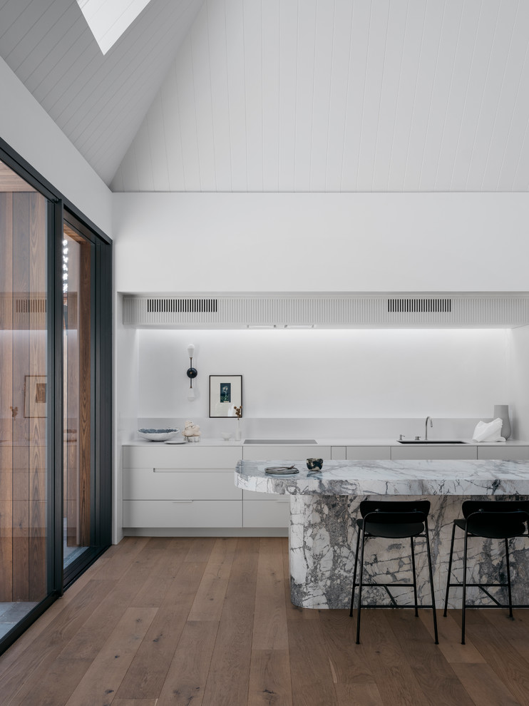 This is an example of a large contemporary single-wall eat-in kitchen in Sydney with an undermount sink, flat-panel cabinets, white cabinets, granite benchtops, white splashback, white appliances, light hardwood floors, with island, brown floor and white benchtop.