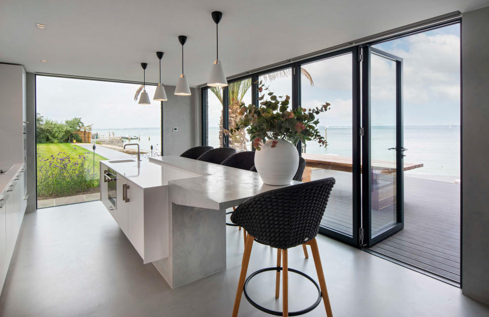Photo of a large beach style galley open plan kitchen in Other with a drop-in sink, flat-panel cabinets, white cabinets, quartz benchtops, panelled appliances, concrete floors, with island, grey floor and white benchtop.