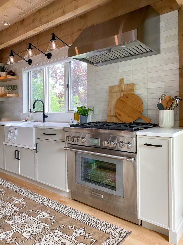 Photo of a transitional l-shaped kitchen in Boston with a farmhouse sink, shaker cabinets, grey cabinets, quartz benchtops, white splashback, ceramic splashback, panelled appliances, light hardwood floors, with island, white benchtop and exposed beam.