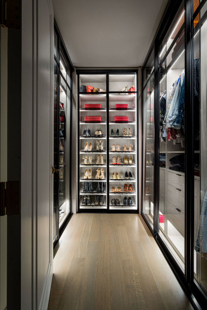 Design ideas for a mid-sized women's walk-in wardrobe in New York with glass-front cabinets, grey cabinets, light hardwood floors and beige floor.