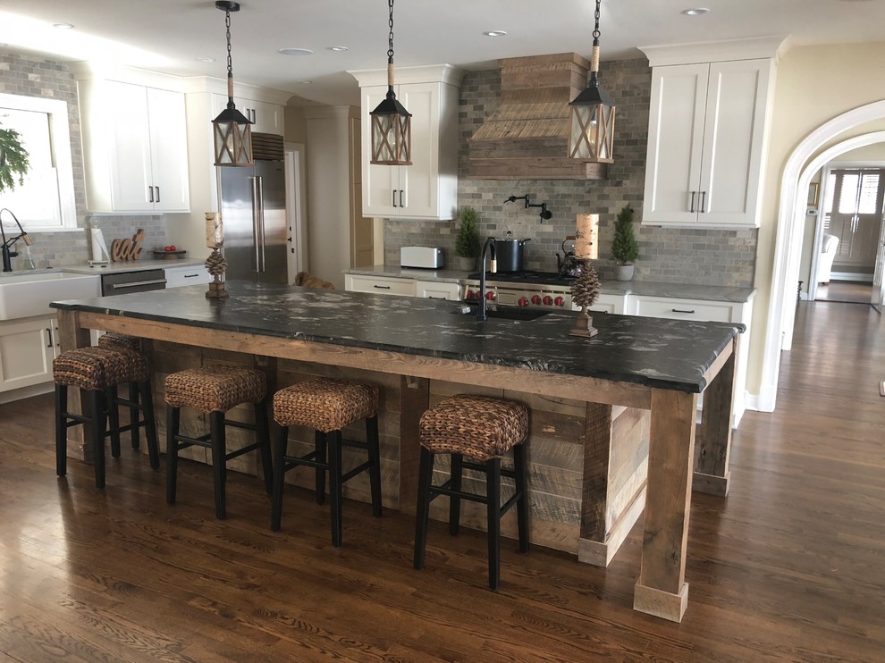 Mid-sized country l-shaped open plan kitchen in New York with a farmhouse sink, shaker cabinets, white cabinets, granite benchtops, grey splashback, stainless steel appliances, medium hardwood floors, with island and brown floor.