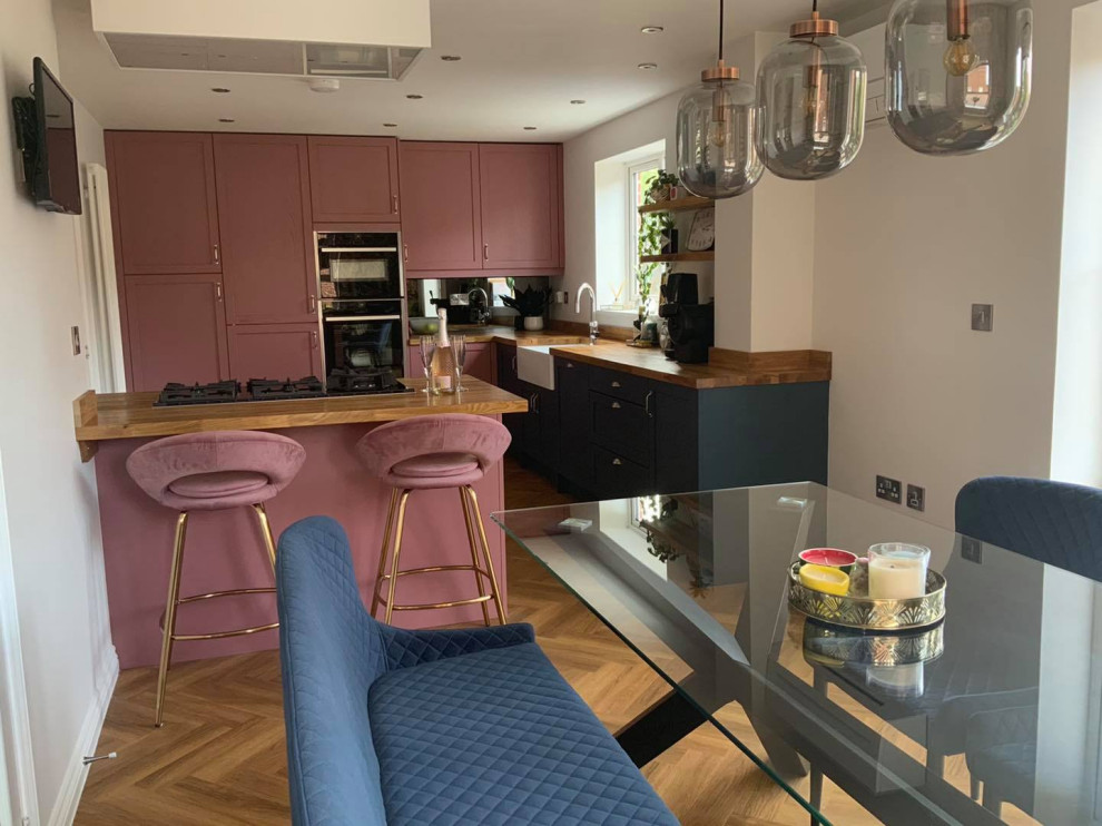 Mid-sized eclectic l-shaped vinyl floor and brown floor eat-in kitchen photo in Buckinghamshire with a farmhouse sink, shaker cabinets, pink cabinets, wood countertops, metallic backsplash, mirror backsplash, paneled appliances, a peninsula and brown countertops