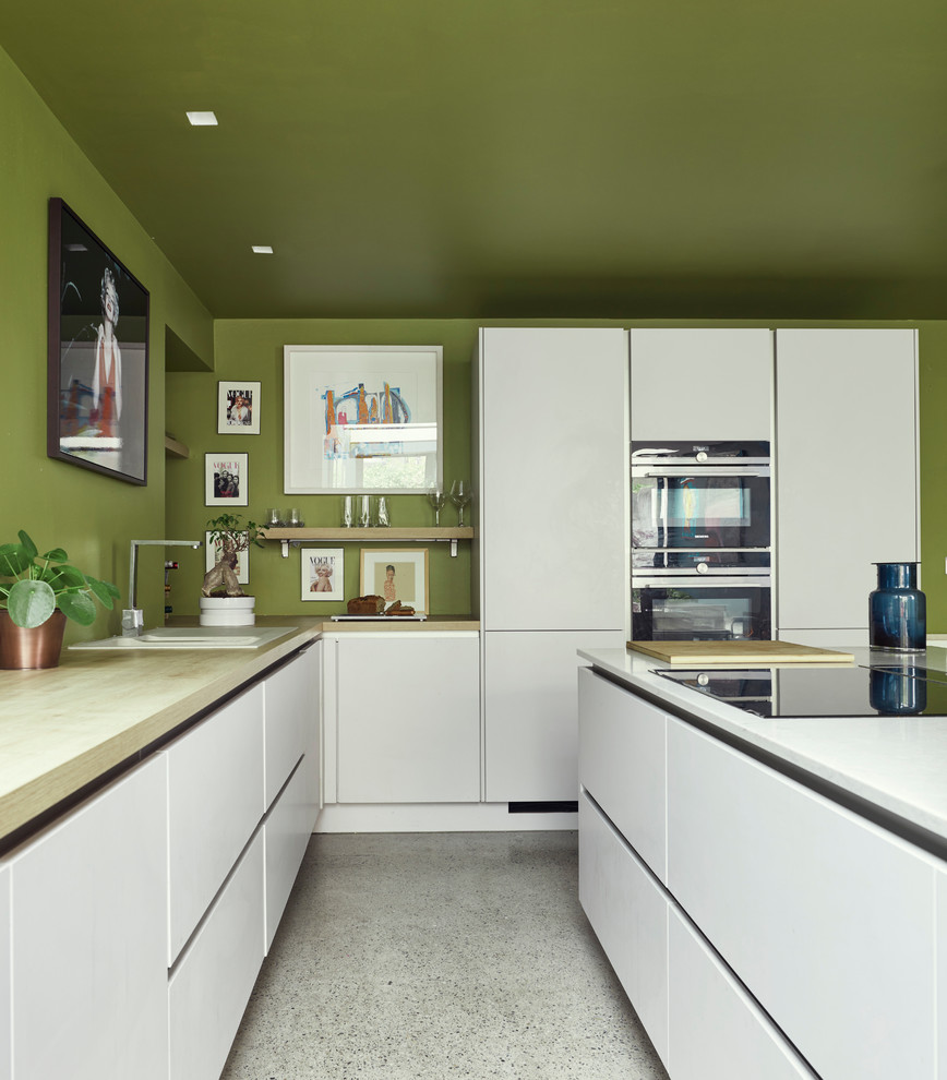 Design ideas for a mid-sized contemporary l-shaped kitchen in Dublin with white cabinets, wood benchtops, with island, white floor, a drop-in sink, flat-panel cabinets, green splashback, black appliances and terrazzo floors.