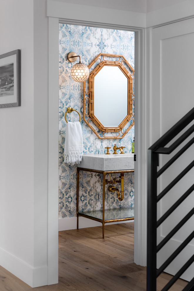 This is an example of a medium sized coastal cloakroom in Other with white cabinets, a one-piece toilet, multi-coloured walls, medium hardwood flooring, a pedestal sink, marble worktops, beige floors, white worktops, a freestanding vanity unit and wallpapered walls.