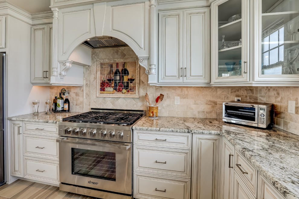 This is an example of a large beach style l-shaped open plan kitchen in New York with an undermount sink, raised-panel cabinets, beige cabinets, granite benchtops, beige splashback, travertine splashback, stainless steel appliances, porcelain floors, with island, beige floor and beige benchtop.