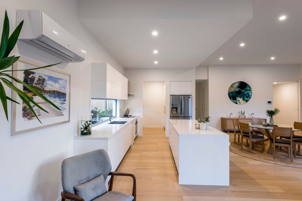 Inspiration for a mid-sized tropical galley open plan kitchen in Gold Coast - Tweed with a double-bowl sink, flat-panel cabinets, white cabinets, quartz benchtops, white splashback, glass sheet splashback, stainless steel appliances, light hardwood floors, with island, brown floor and white benchtop.