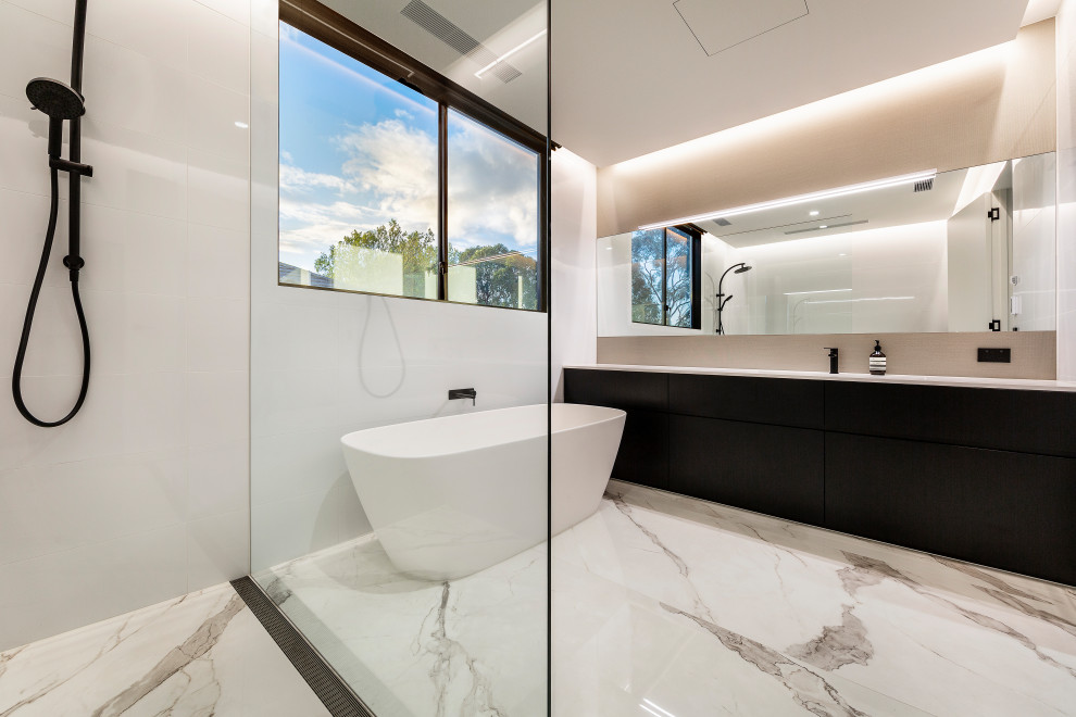 Photo of a large contemporary master bathroom in Melbourne with raised-panel cabinets, black cabinets, a freestanding tub, an open shower, a wall-mount toilet, beige tile, porcelain tile, white walls, marble floors, a drop-in sink, marble benchtops, white floor, an open shower and white benchtops.