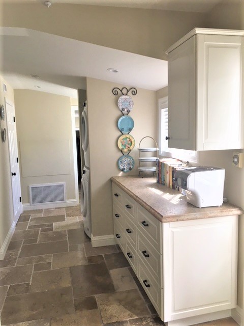 Mid-sized traditional galley utility room in Los Angeles with white cabinets, granite benchtops, beige walls, travertine floors, a stacked washer and dryer, beige floor, beige benchtop and raised-panel cabinets.