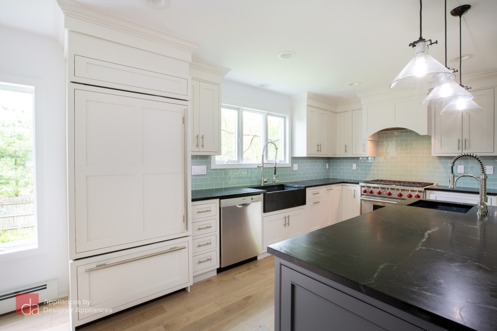 This is an example of a large transitional l-shaped open plan kitchen in New York with a farmhouse sink, shaker cabinets, white cabinets, quartz benchtops, green splashback, subway tile splashback, panelled appliances, light hardwood floors, with island, brown floor and black benchtop.