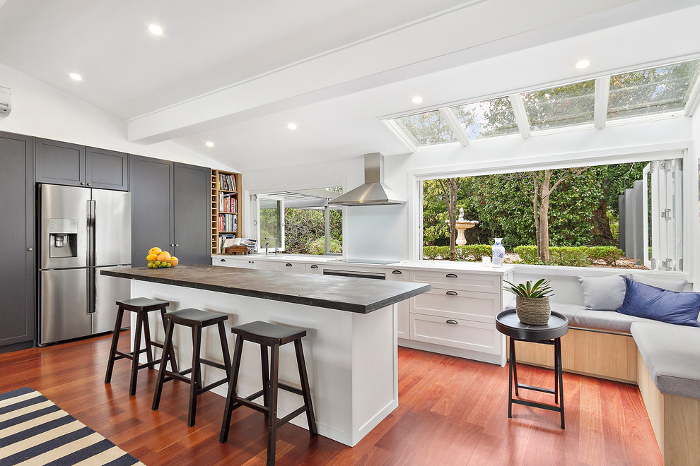 This is an example of an expansive contemporary l-shaped open plan kitchen in Sydney with brown floor, shaker cabinets, with island, an integrated sink, white cabinets, zinc benchtops, white splashback, glass sheet splashback, black appliances, light hardwood floors and grey benchtop.