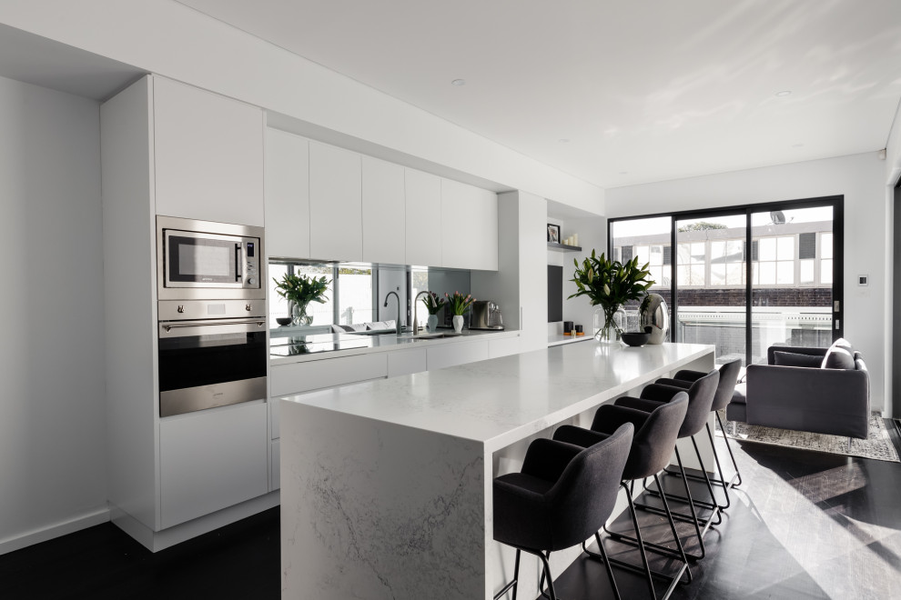 Design ideas for a mid-sized contemporary open plan kitchen in London with a drop-in sink, glass-front cabinets, white cabinets, quartzite benchtops, metallic splashback, glass sheet splashback, stainless steel appliances, dark hardwood floors, with island, brown floor and white benchtop.