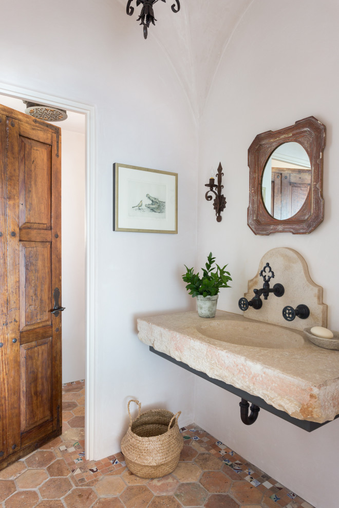 This is an example of a mid-sized mediterranean powder room in Los Angeles with a one-piece toilet, beige walls, terra-cotta floors, a wall-mount sink, limestone benchtops, red floor, beige benchtops, a floating vanity and vaulted.