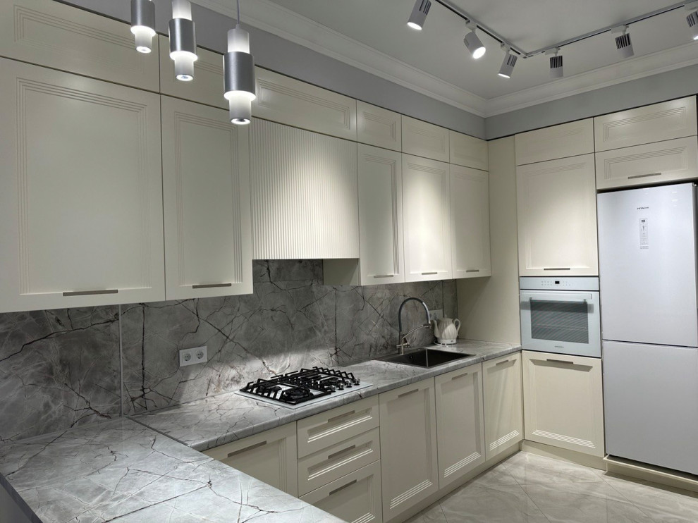 На фото: отдельная, угловая, серо-белая кухня среднего размера: освещение в стиле неоклассика (современная классика) с врезной мойкой, белыми фасадами, серым фартуком, полуостровом и серой столешницей