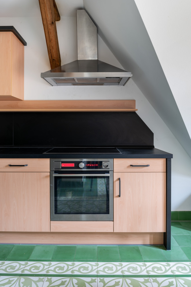 Photo of a large country galley eat-in kitchen in Other with an integrated sink, flat-panel cabinets, light wood cabinets, solid surface benchtops, black splashback, slate splashback, stainless steel appliances, porcelain floors, no island, green floor and black benchtop.