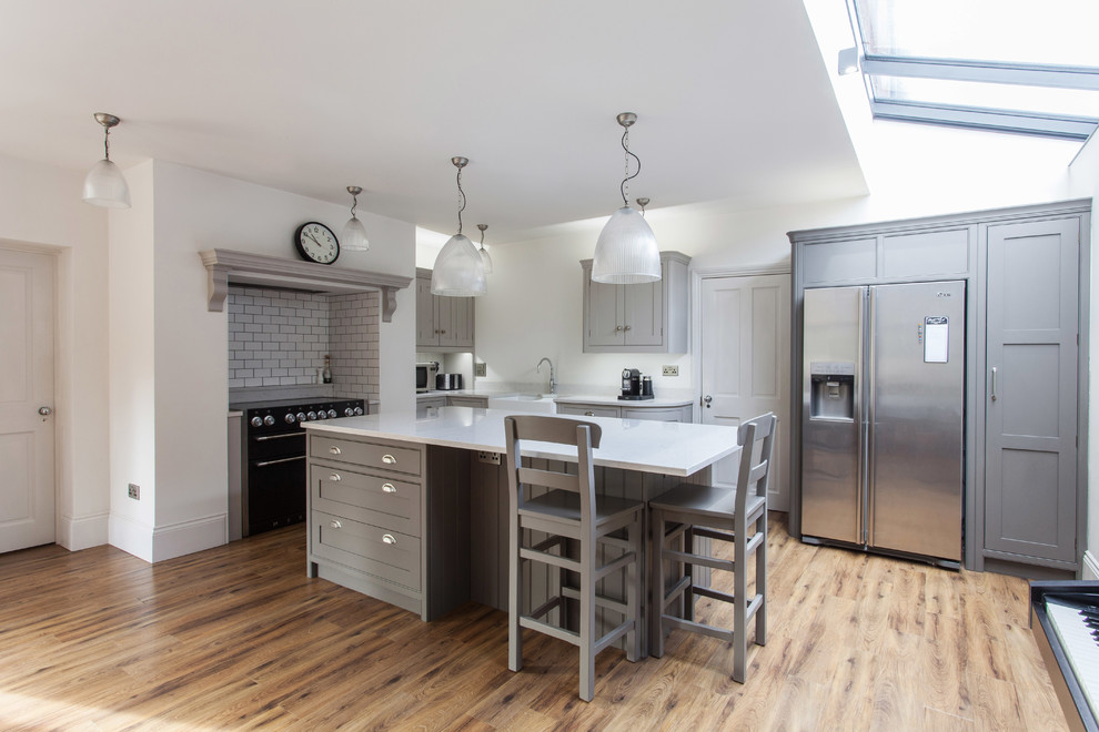 Inspiration for a mid-sized transitional l-shaped eat-in kitchen in Kent with a farmhouse sink, beaded inset cabinets, grey cabinets, quartzite benchtops, white splashback, subway tile splashback, black appliances, light hardwood floors, with island and beige floor.