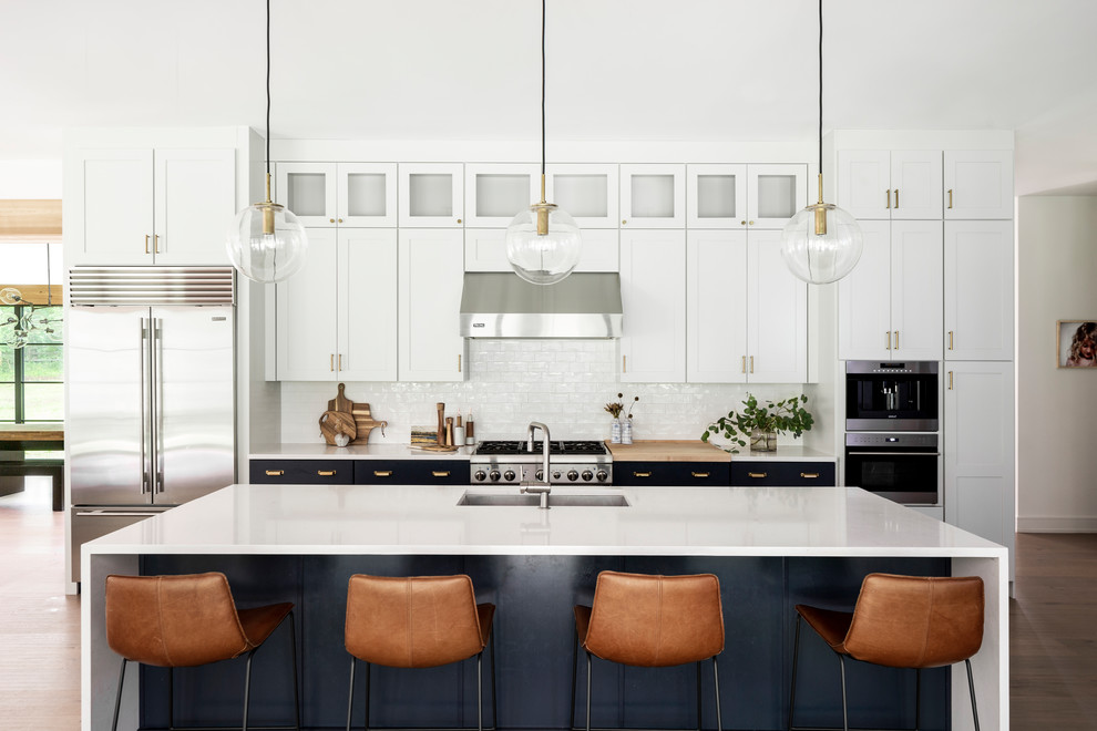 Inspiration for a transitional kitchen in New York with an undermount sink, shaker cabinets, white cabinets, white splashback, subway tile splashback, stainless steel appliances, with island, brown floor and white benchtop.