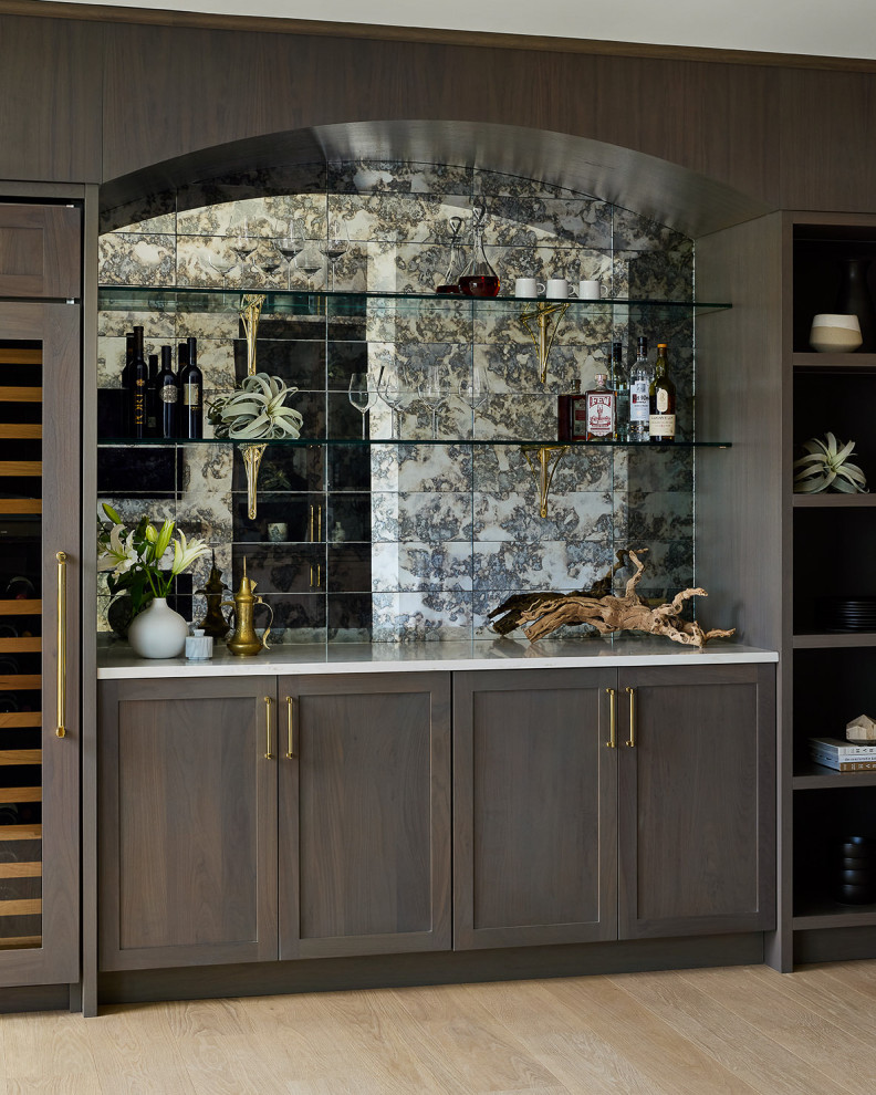 This is an example of a small transitional single-wall home bar in Chicago with shaker cabinets, brown cabinets, quartz benchtops, glass tile splashback, light hardwood floors and white benchtop.
