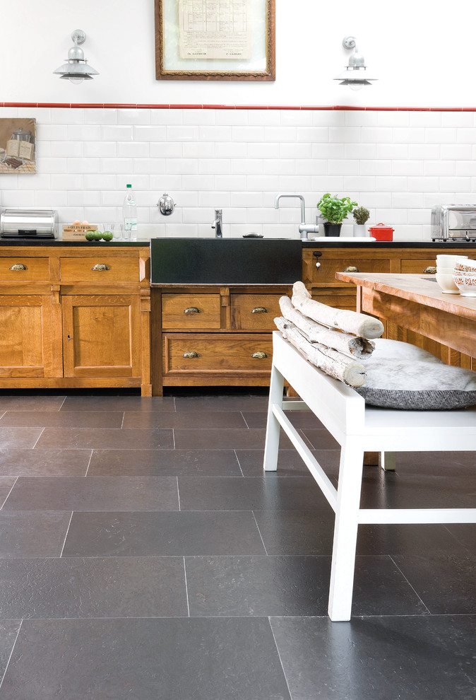 This is an example of a mid-sized country single-wall separate kitchen in Other with a farmhouse sink, raised-panel cabinets, medium wood cabinets, white splashback, subway tile splashback, slate floors, no island, black floor and black benchtop.