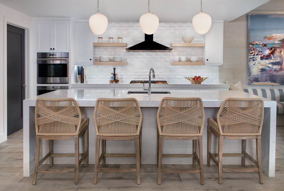 Design ideas for a beach style galley open plan kitchen in Cleveland with an undermount sink, shaker cabinets, white cabinets, white splashback, stainless steel appliances, light hardwood floors, with island, grey floor and grey benchtop.