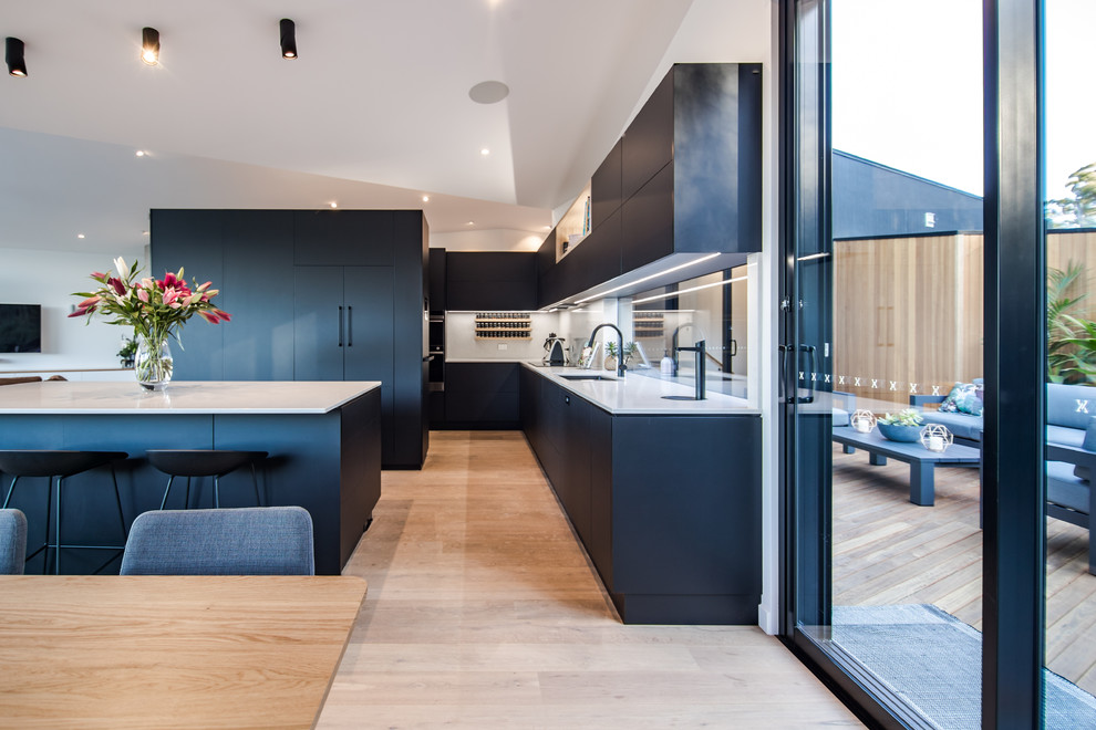 This is an example of a contemporary eat-in kitchen in Hobart with an undermount sink, flat-panel cabinets, black cabinets, white splashback, panelled appliances, light hardwood floors, with island, beige floor and white benchtop.