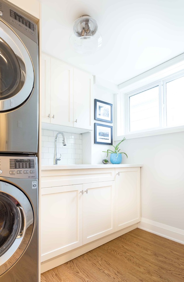 Photo of a mid-sized midcentury galley dedicated laundry room in Toronto with an undermount sink, shaker cabinets, white cabinets, solid surface benchtops, grey walls, light hardwood floors and a stacked washer and dryer.