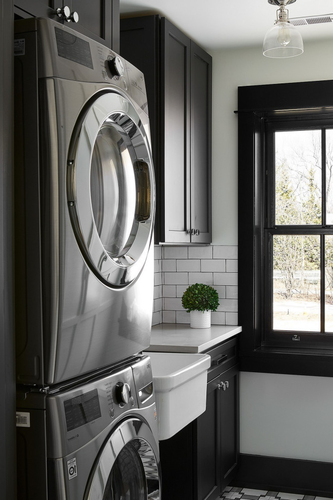 Inspiration for a small transitional single-wall dedicated laundry room in Orlando with a farmhouse sink, shaker cabinets, black cabinets, solid surface benchtops, white walls, a stacked washer and dryer, multi-coloured floor and white benchtop.