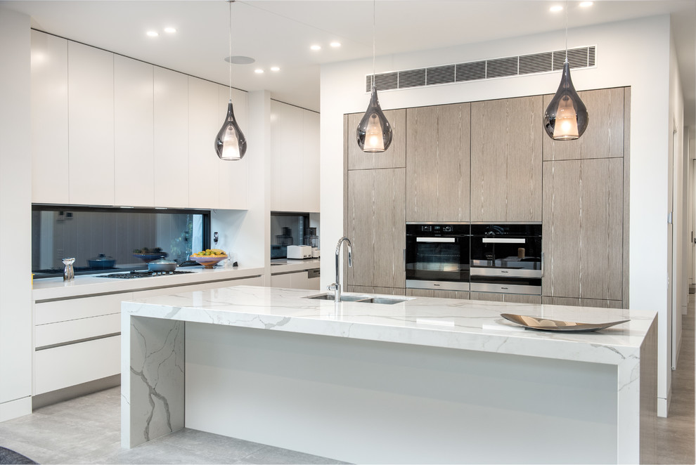 This is an example of a contemporary galley kitchen in Melbourne with a double-bowl sink, flat-panel cabinets, light wood cabinets, marble benchtops, glass sheet splashback, stainless steel appliances, light hardwood floors and with island.