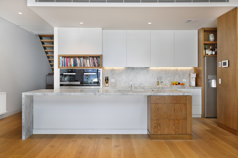 Photo of a large contemporary u-shaped kitchen pantry in Melbourne with an undermount sink, raised-panel cabinets, white cabinets, quartz benchtops, grey splashback, stone slab splashback, stainless steel appliances, light hardwood floors, with island and beige floor.