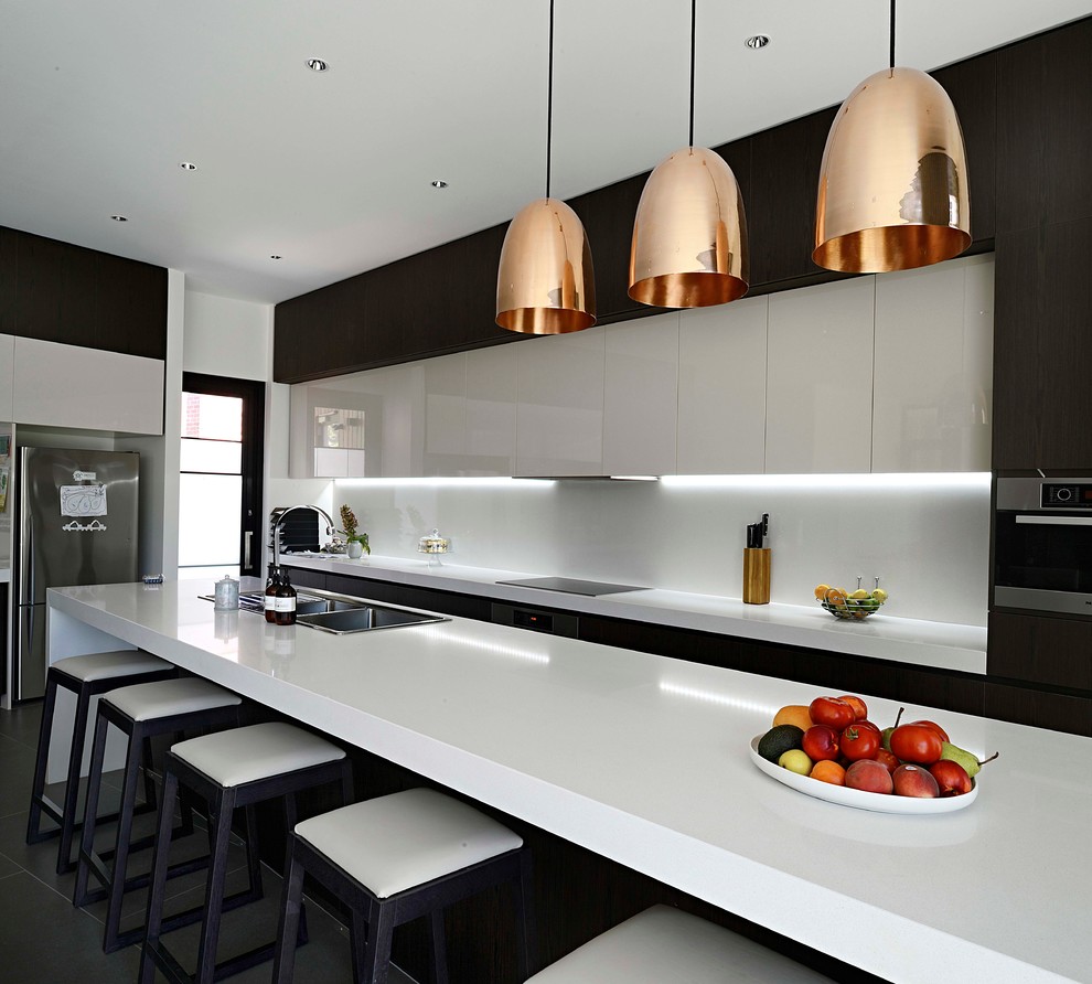 This is an example of a large contemporary galley open plan kitchen in Melbourne with a double-bowl sink, flat-panel cabinets, dark wood cabinets, white splashback, stainless steel appliances, with island, grey floor and white benchtop.