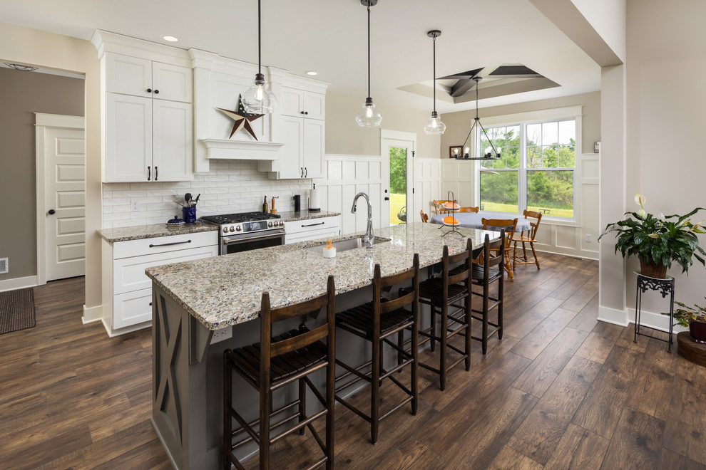 Mid-sized traditional galley eat-in kitchen in Grand Rapids with a farmhouse sink, recessed-panel cabinets, white cabinets, granite benchtops, white splashback, subway tile splashback, stainless steel appliances, laminate floors, with island, brown floor and beige benchtop.