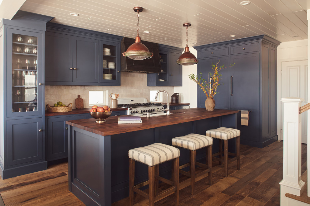Enclosed kitchen - large coastal l-shaped medium tone wood floor enclosed kitchen idea in Orange County with a farmhouse sink, recessed-panel cabinets, blue cabinets, wood countertops, beige backsplash, stone tile backsplash, stainless steel appliances and an island